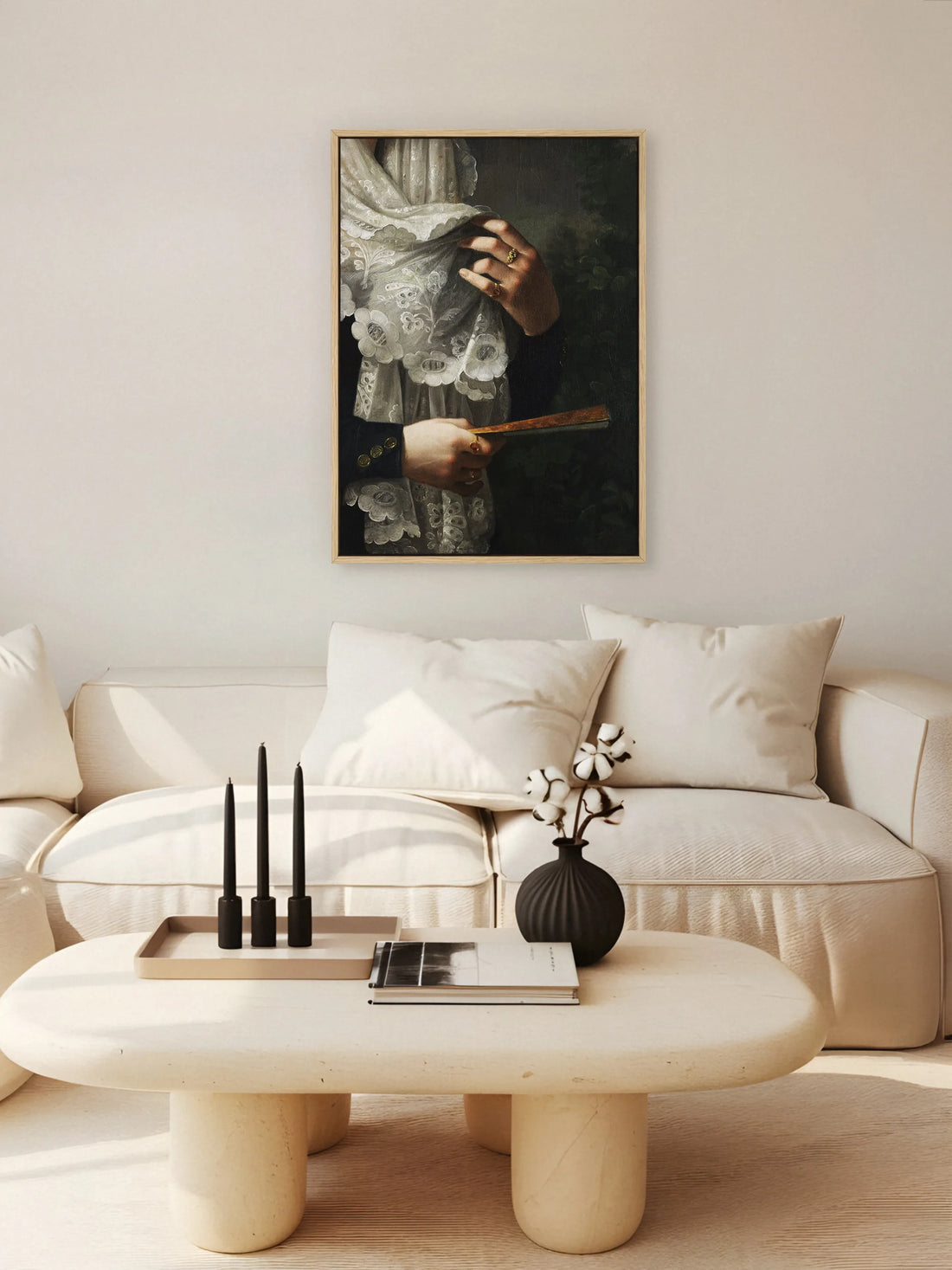 Leinwandbild mit Spitze und Fächer im naturfarbenen Rahmen über einem cremefarbenen Sofa mit Couchtisch aus Stein, schwarzen Kerzen und Baumwollzweigen