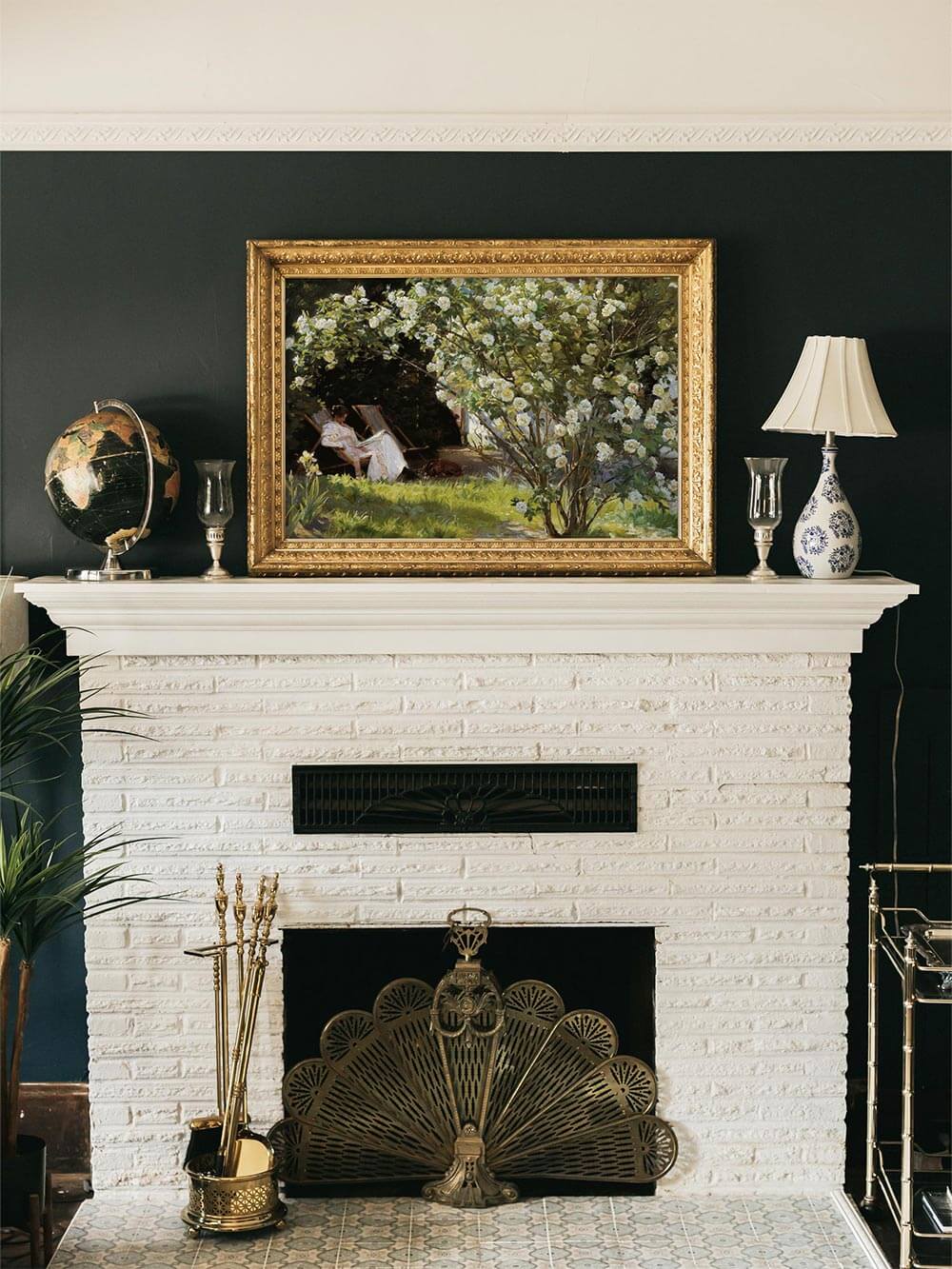 Elegantes Vintage-Wandbild über einem weißen Kamin mit goldverzierter Rahmenoptik. Das impressionistische Gartenmotiv zeigt eine ruhige Szene mit einer lesenden Frau unter einem blühenden Rosenbusch. Perfekt für klassische Wohnräume und stilvolle Wandgestaltung.