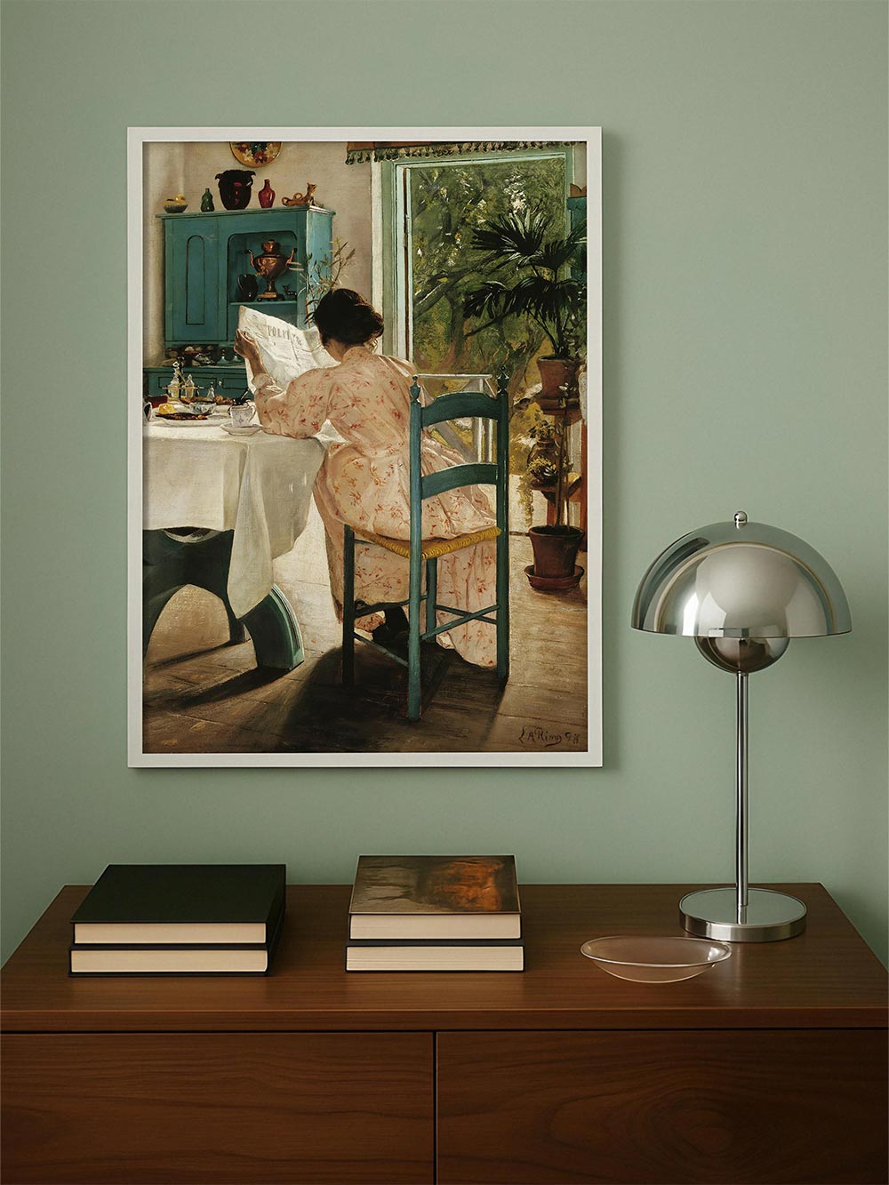 Gerahmter Kunstdruck einer Frau im geblümten Kleid beim Lesen am gedeckten Tisch vor grünem Fenster, präsentiert über einer Kommode in ruhigem Wohnraum. Klassisches Wandbild im Vintage-Stil für elegante Wohn- und Leseecken.