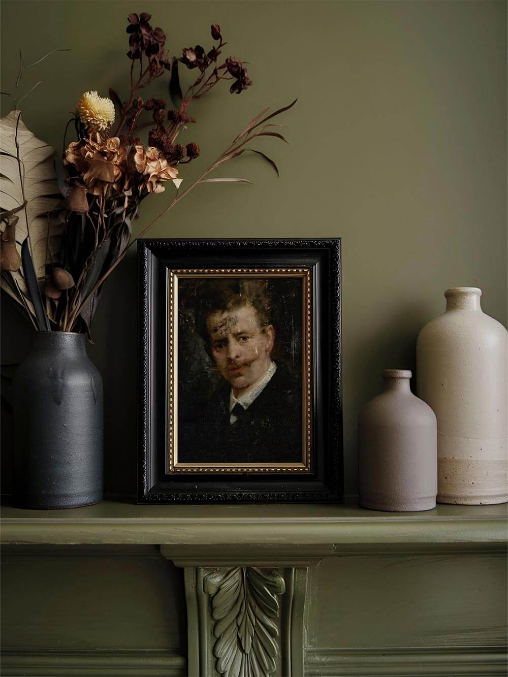 Ein Männerporträt-Kunstdruck im schwarzen Rahmen steht auf einem Kaminsims neben Trockenblumen und Keramikvasen. Das dunkle Bild fügt sich harmonisch ins nostalgische Interieur.