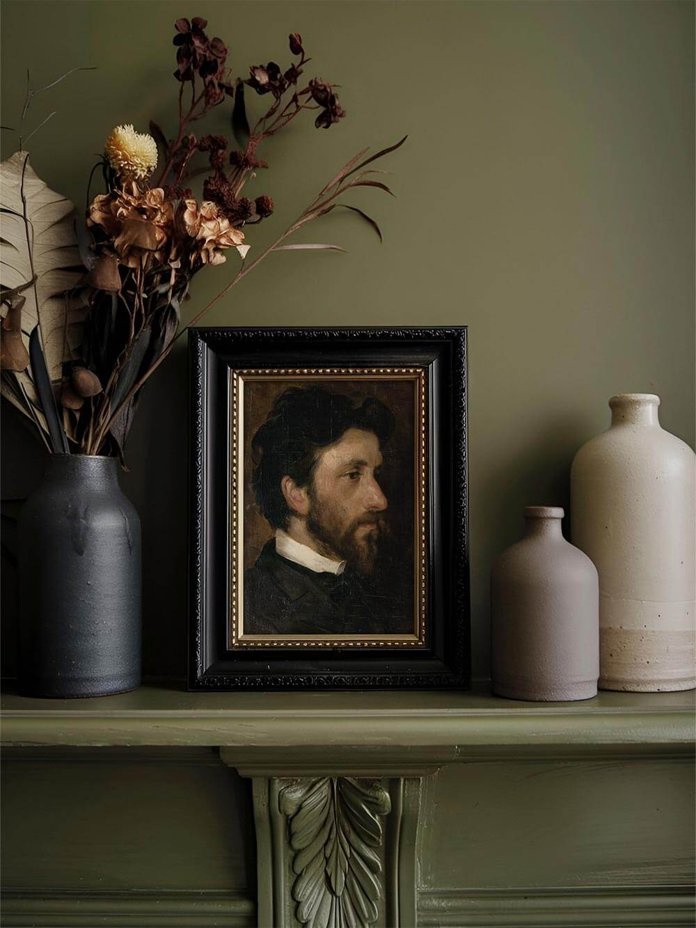 Der Männerporträt-Kunstdruck im schwarzen Rahmen steht auf einem Kaminsims neben Trockenblumen und Keramikvasen. Das dunkle Bild fügt sich harmonisch ins nostalgische Interieur.
