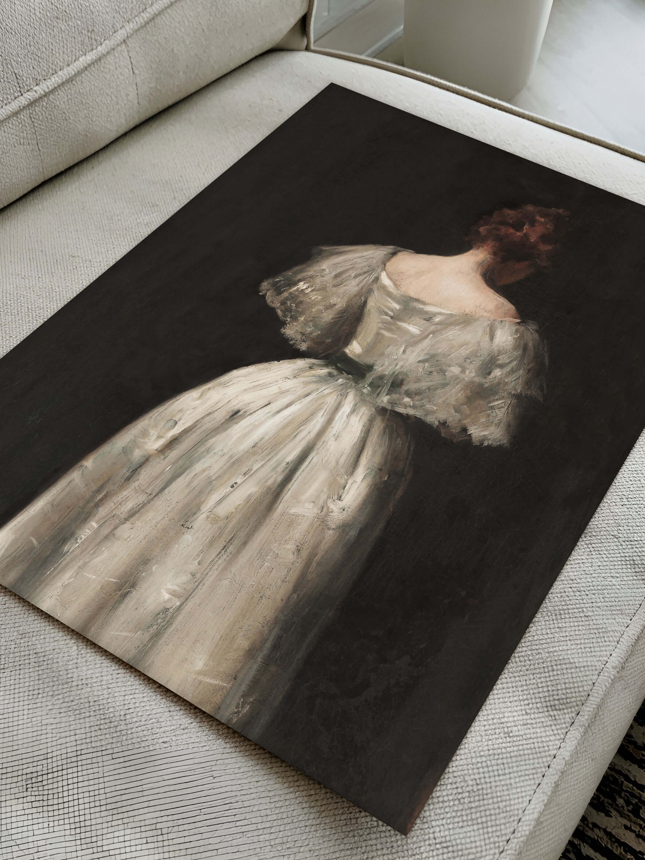 Kunstdruck eines dunklen Gemäldes, der den Rücken einer Frau in einem weißen Kleid zeigt. Das Poster liegt auf einer modernen Couch.