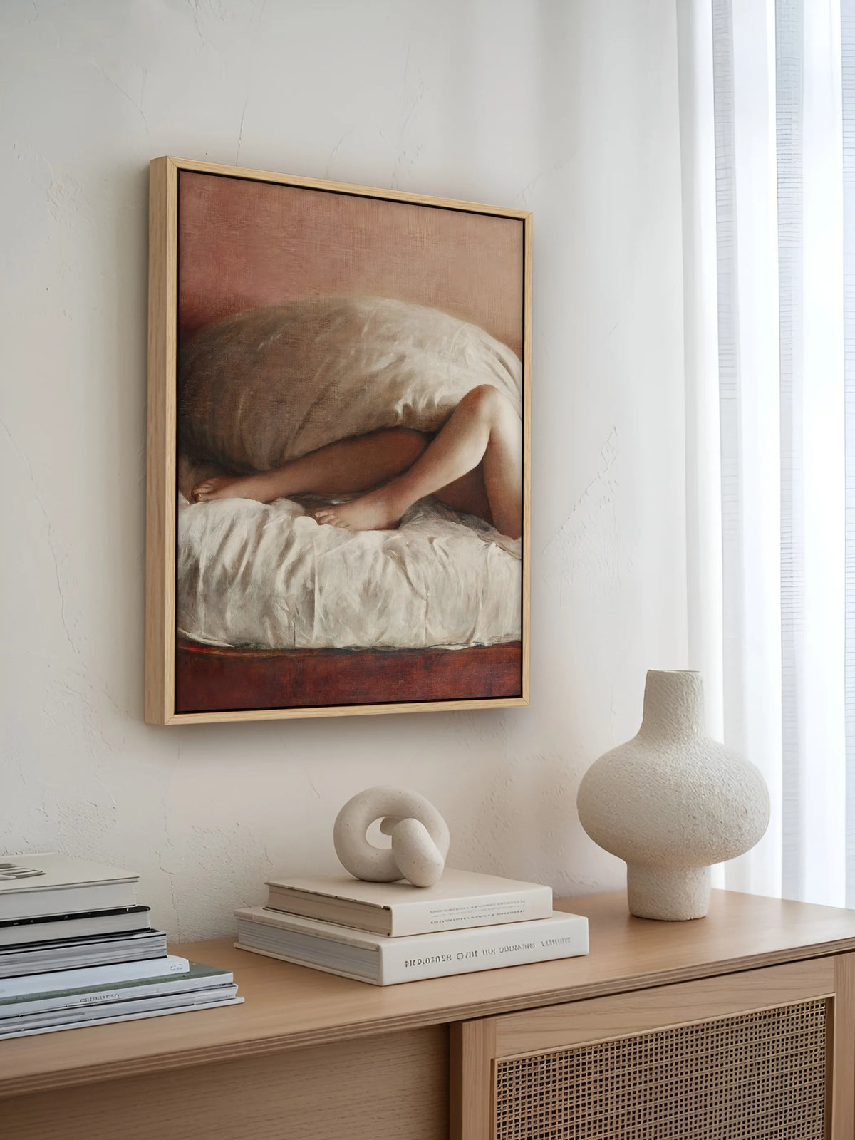 Leinwandbild mit verhülltem Akt im naturfarbenen Rahmen über einem Sideboard mit Rattanfronten, hellen Büchern und Vase aus Keramik am Fenster