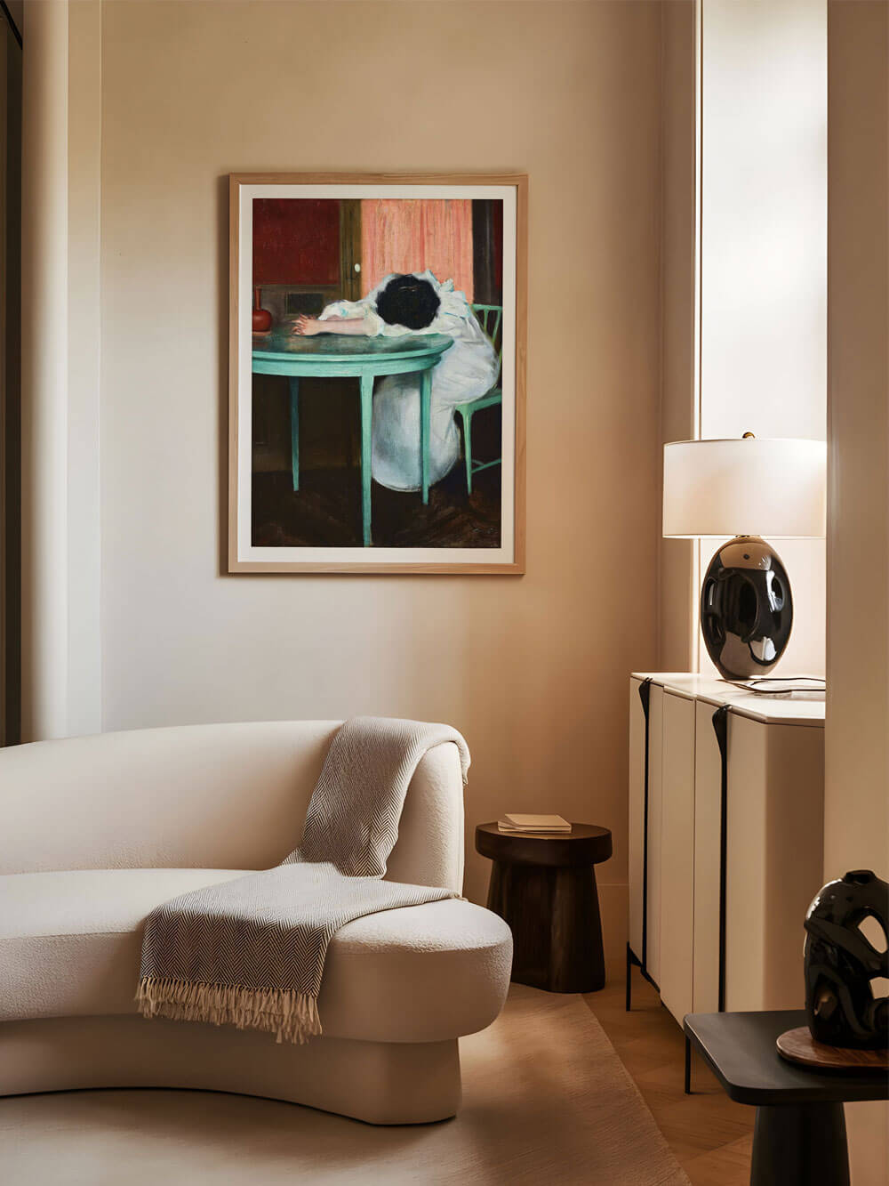 Vintage-Kunstdruck einer Frau, die mit dem Kopf auf einem Tisch ruht, stilvoll eingerahmt und an einer beigefarbenen Wohnzimmerwand über einem geschwungenen, cremefarbenen Sofa aufgehängt.