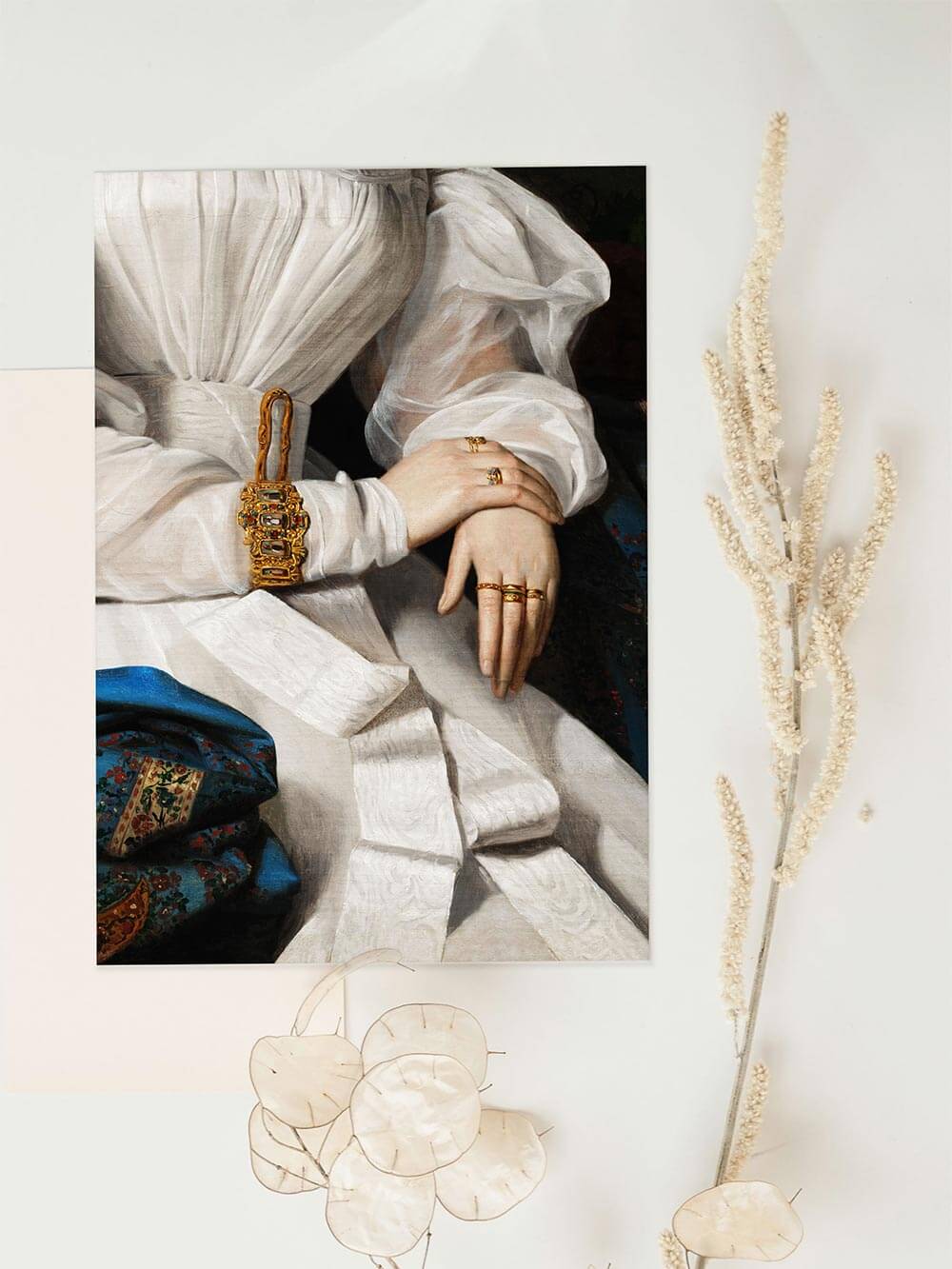 Kunstdruck zeigt gefalteten weißen Stoff und goldene Schmuckelemente auf hellem Untergrund mit Trockenblumen. Ästhetische Komposition mit Fokus auf Textur, Licht und historische Eleganz.

