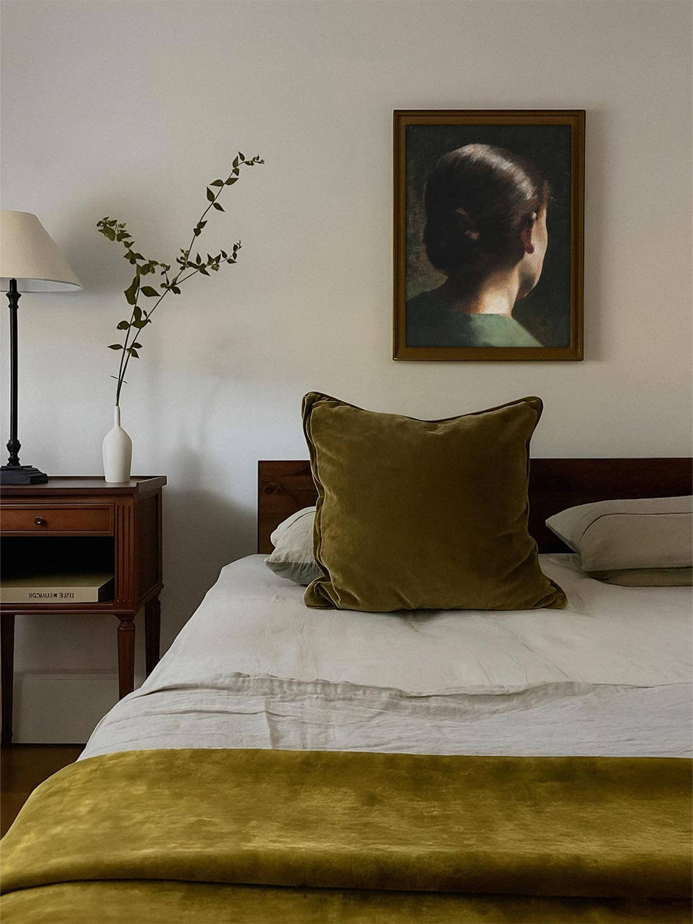 Gerahmtes Porträt einer Frau von hinten über einem Bett mit goldgrünen Textilien und minimalistischer Dekoration. Sanfte, ruhige Stimmung – idealer Kunstdruck für elegante und harmonische Schlafzimmergestaltung.