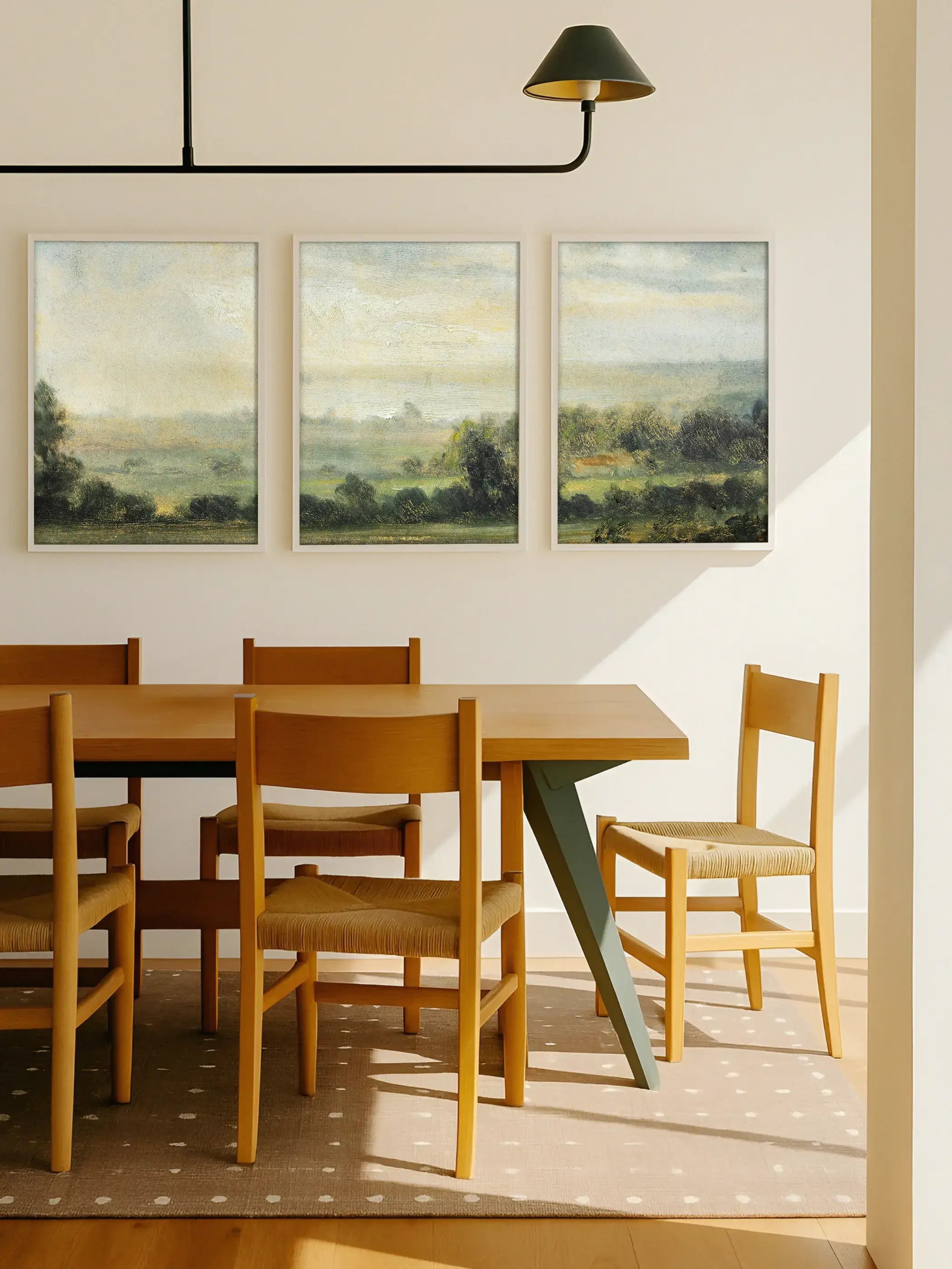 Drei gerahmte Landschafts-Poster mit ruhiger Hügellandschaft hängen über einem Holztisch im hellen Esszimmer. Die soften Naturtöne ergänzen das warme Interieur und schaffen eine entspannte, stilvolle Galerie-Wand.