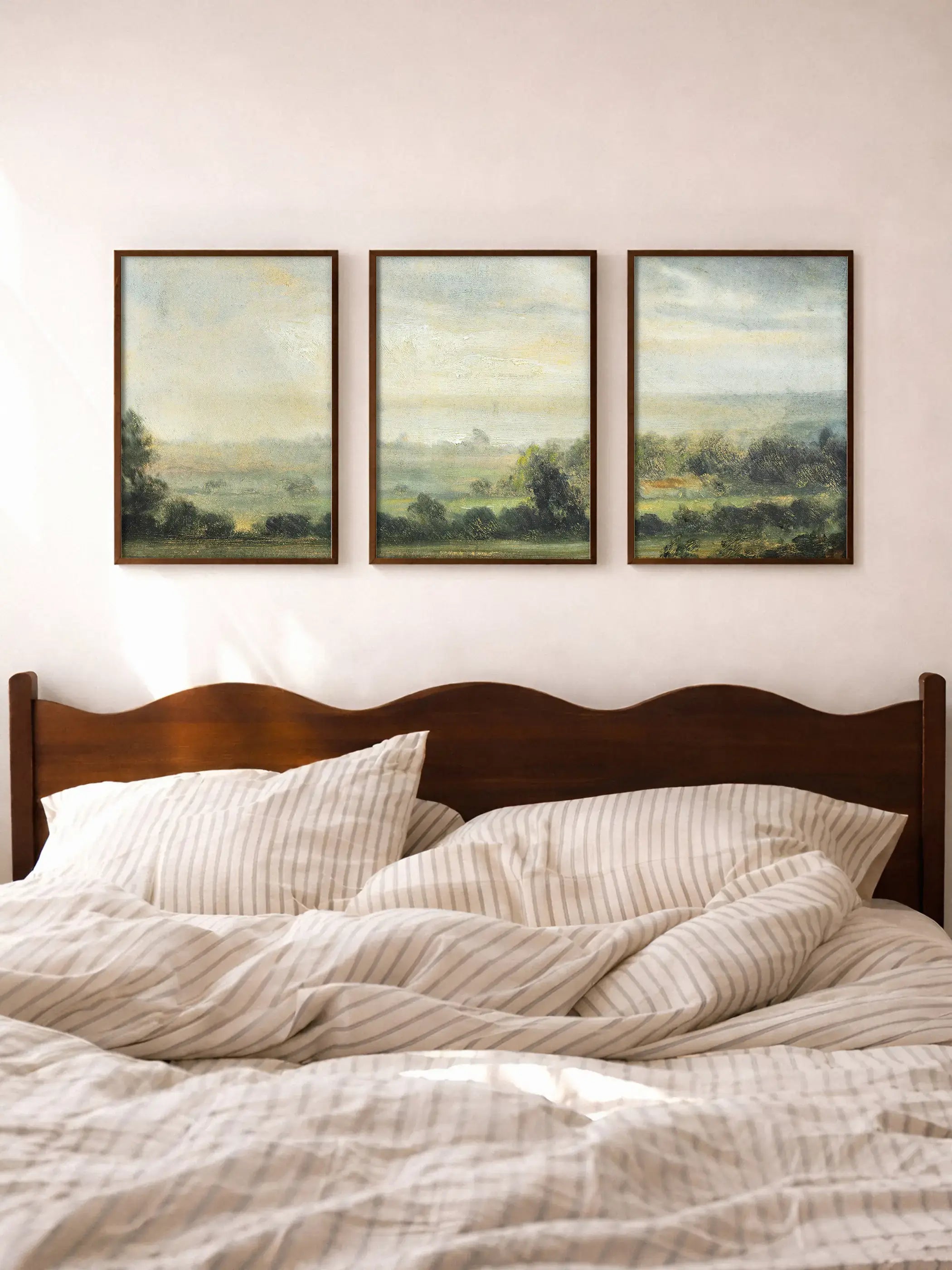 Dreiteiliges Landschaftsbild mit sanften Feldern und Bäumen in gedeckten Grün- und Beigetönen hängt über einem Bett mit heller Bettwäsche. Das ruhige Wandbild bringt eine harmonische und beruhigende Atmosphäre ins Schlafzimmer.