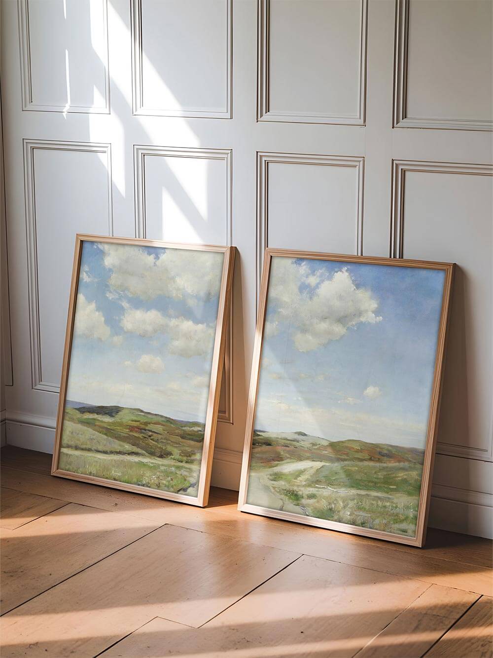 Zwei gerahmte Landschafts-Kunstdrucke lehnen an einer Wand im Sonnenlicht. Die Kombination aus Holzrahmen und pastelligen Tönen vermittelt Wärme, Schlichtheit und natürliche Eleganz im Wabi-Sabi-Stil.