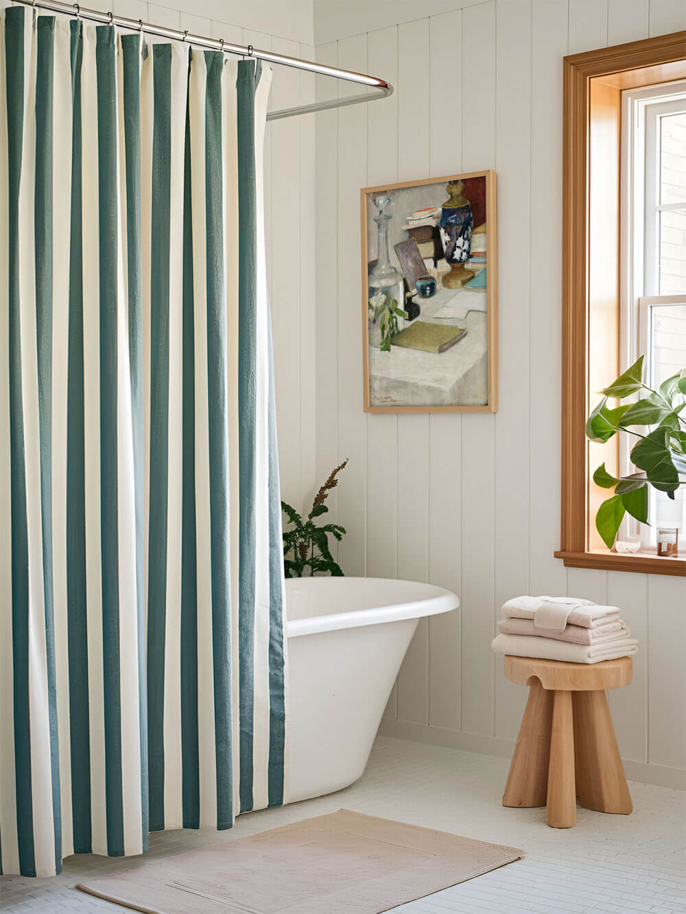 Vintage-Stillleben-Kunstdruck in einem Badezimmer mit weiß-blau gestreiftem Duschvorhang und freistehender Badewanne. Der Kunstdruck hängt an der Wand neben einem Fenster, das Tageslicht hereinlässt und das Bild harmonisch in Szene setzt.