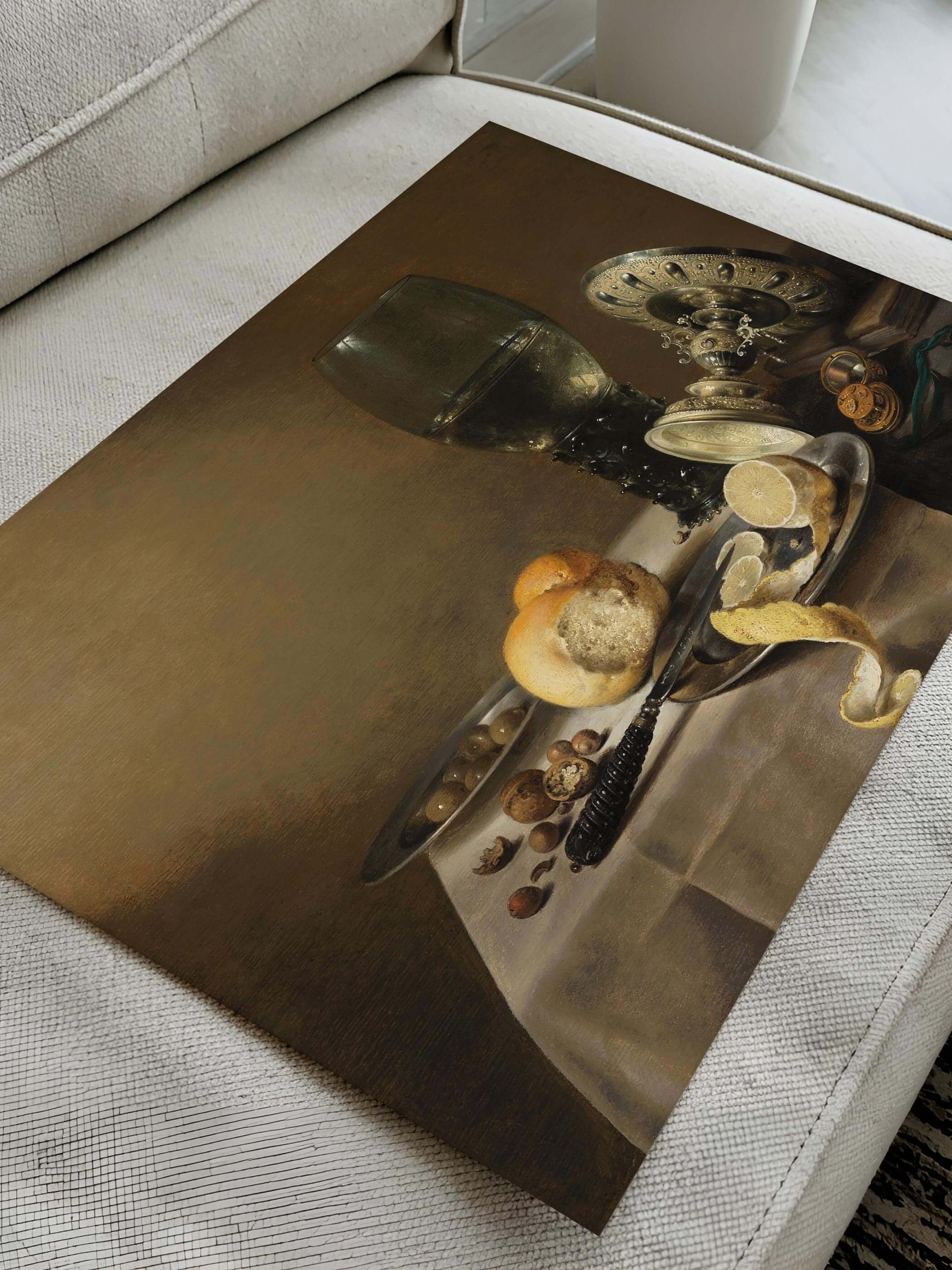 Barockes Stillleben Poster mit Weinglas, Brot und Zitronenschale, gedruckt auf mattem Papier auf einem weißen Sofa liegend. Detaillierter Kunstdruck in einem modernen Wohnraum präsentiert