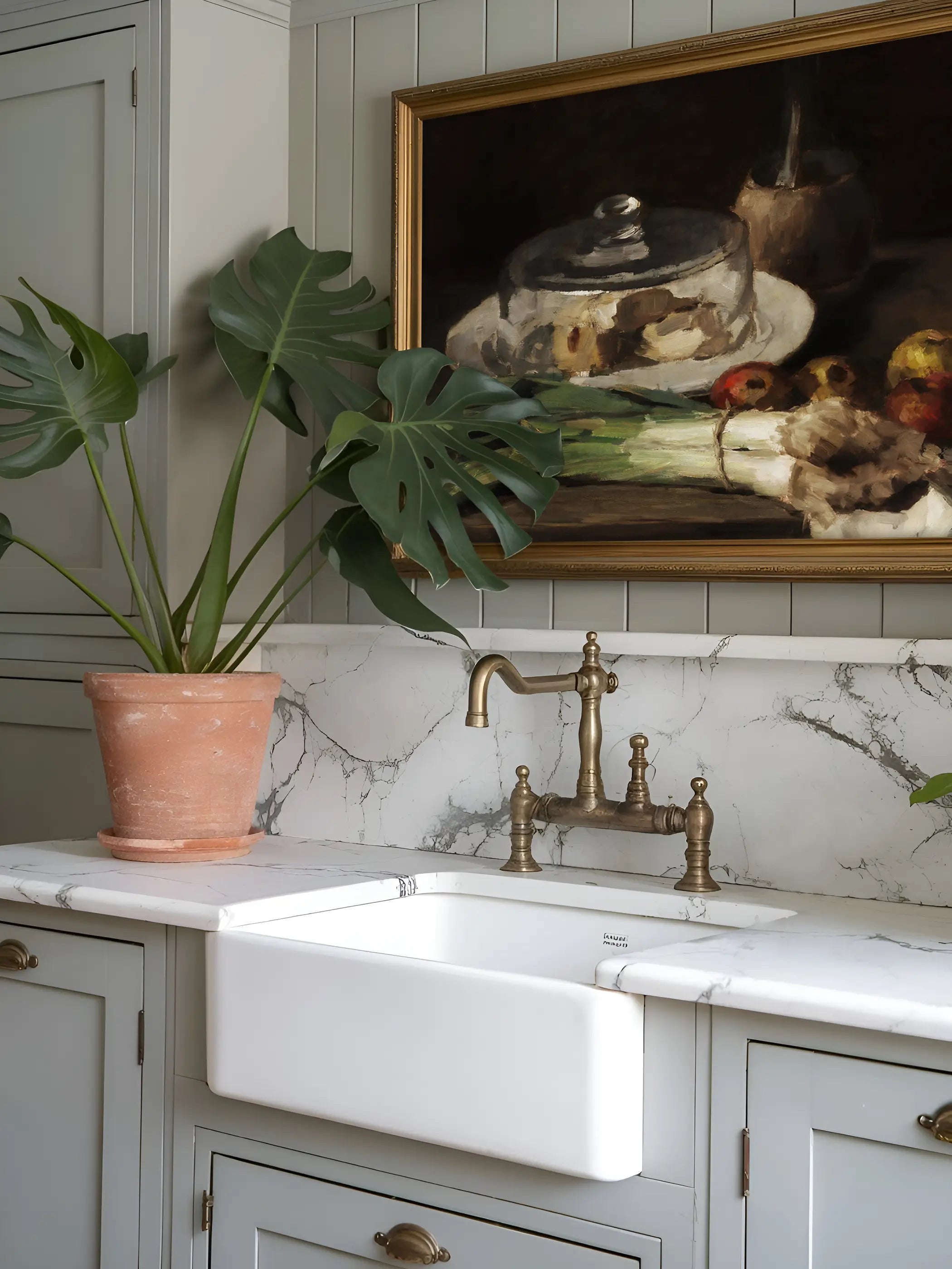 Golden gerahmter Kunstdruck mit Käseglocke, Krug und Lauch hängt über einer weißen Landhausspüle mit Marmorrückwand und Messingarmatur. Der warme Vintage Küchenprint fügt sich neben der Monstera-Pflanze harmonisch in die hellgraue, klassische Küchengestaltung ein.