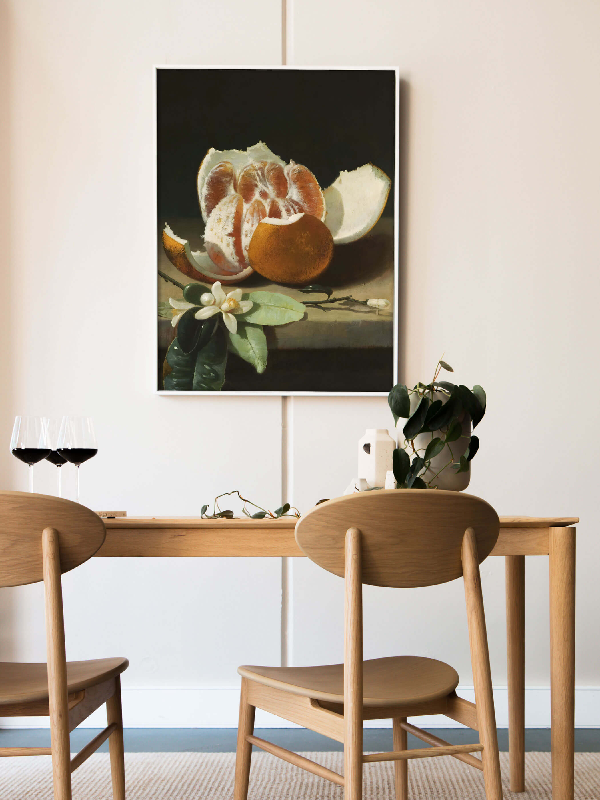 Weiß gerahmter Kunstdruck mit Orangenstillleben lehnt an der Wand über einem Holztisch mit Rotweingläsern und einer grünen Pflanze. Das dunkle, warme Motiv setzt einen eleganten Akzent im hellen Esszimmer.