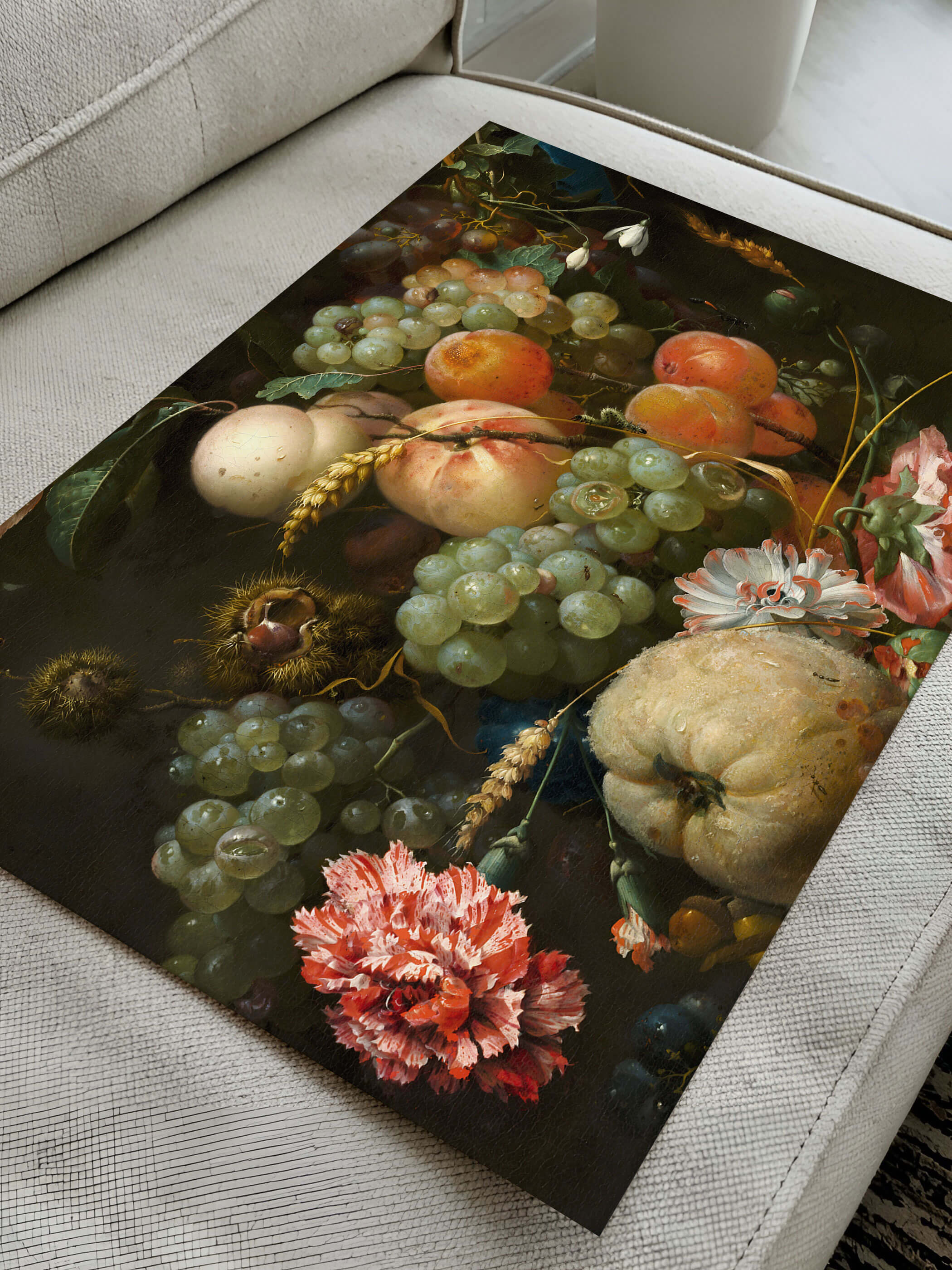 Kunstdruck mit barocker Frucht- und Blumengirlande liegt diagonal auf einem cremefarbenen Sofa. Die Nahaufnahme zeigt die Papierqualität und die satten Farben der Trauben, Pfirsiche und Nelken im Detail.