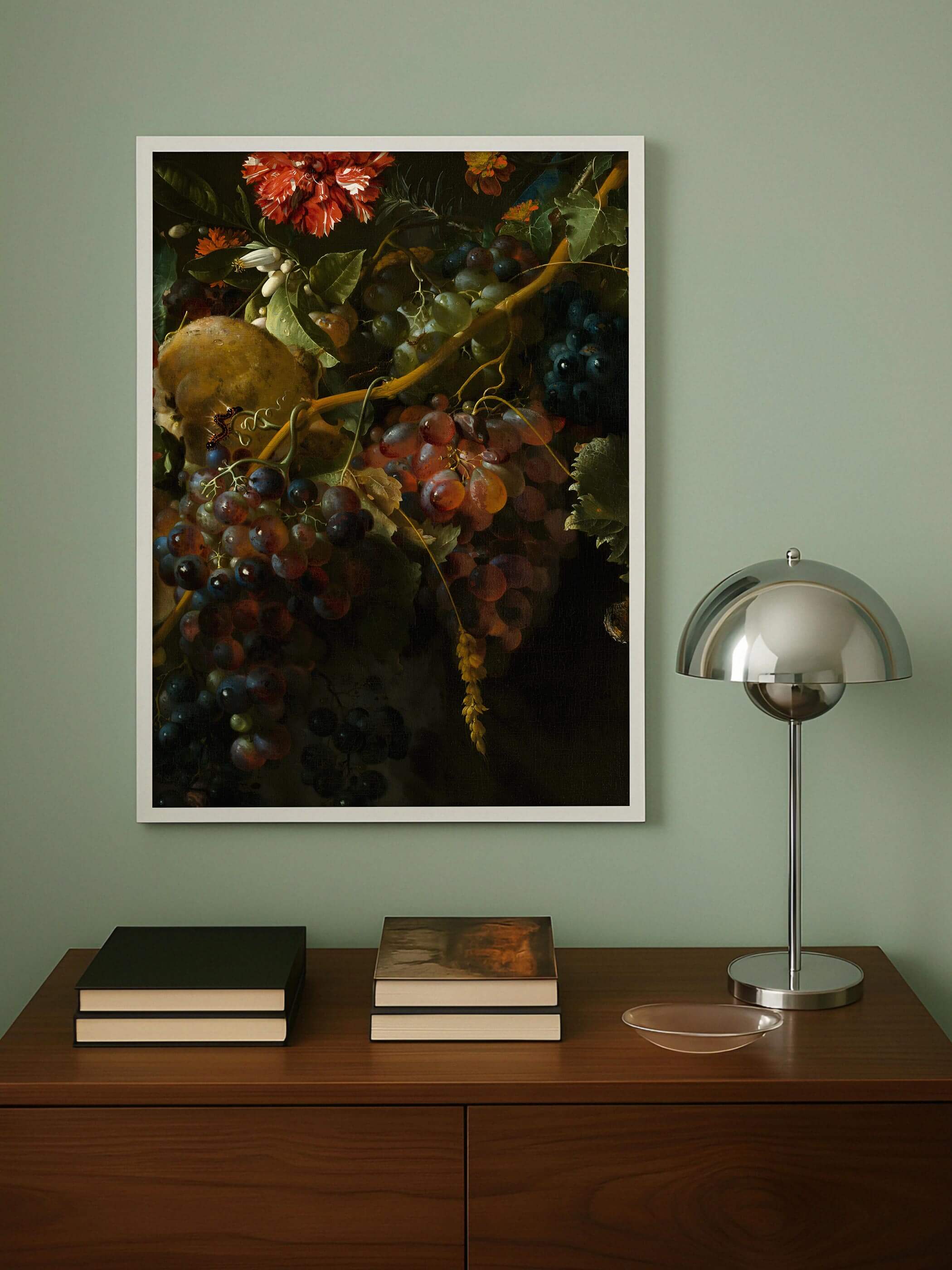Weiß gerahmter Kunstdruck mit dunklem Trauben-Stillleben hängt an einer salbeigrünen Wand über einer Holzkommode mit Büchern und einer Chromlampe. Der dramatische Hell-Dunkel-Kontrast des Motivs bildet einen kräftigen Akzent im ruhigen Raum.