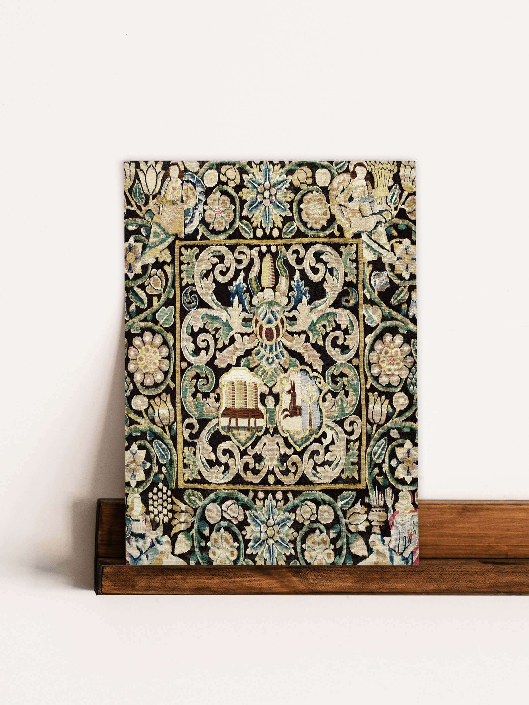 Kunstdruck eines antiken Gobelins mit Wappen, der auf einem Holzregal an einer weiße Wand lehnt. Der Druck zeigt detaillierte florale Muster und Wappenmotive.