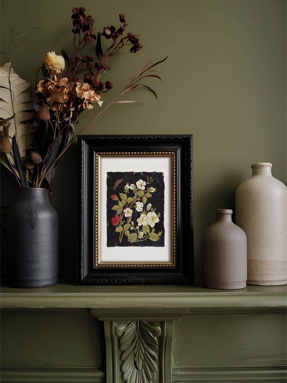 Eleganter Vintage-Kunstdruck mit floraler Stickerei in dunklem Holzrahmen, präsentiert auf einem olivgrünen Kaminsims. Zarte weiße, rosa und rote Blüten auf dunklem Hintergrund erzeugen eine nostalgische Ästhetik – ideal für klassisches oder botanisches Interieur.