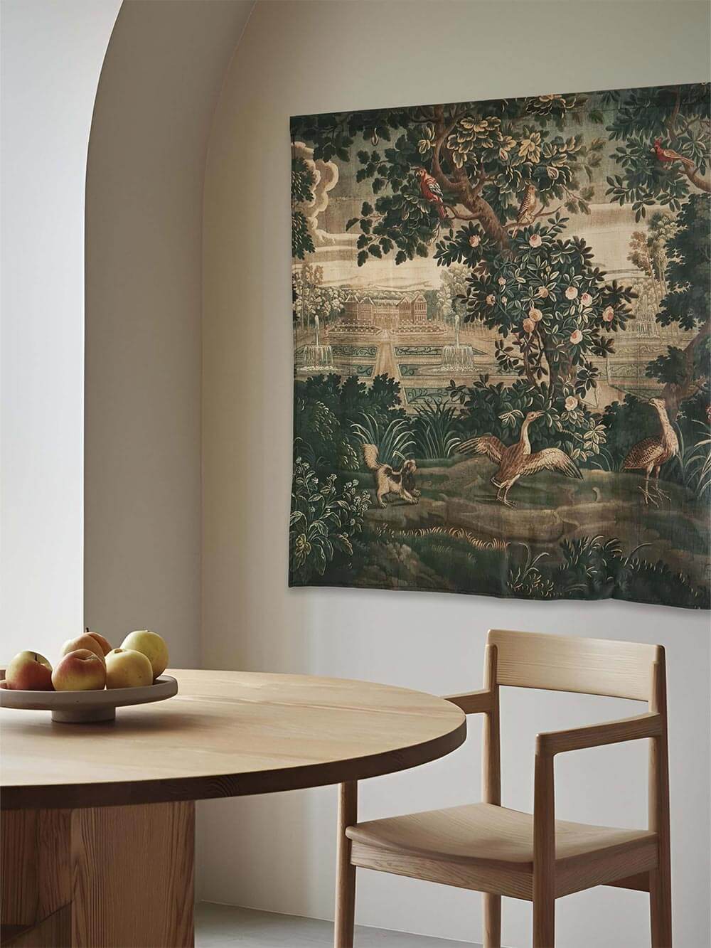 Eleganter Rokoko Wandteppich mit paradiesischer Gartenlandschaft, hängend in einem modernen Esszimmer mit runder Holztischgruppe. Warme Naturtöne betonen die historische Ästhetik des Designs.
