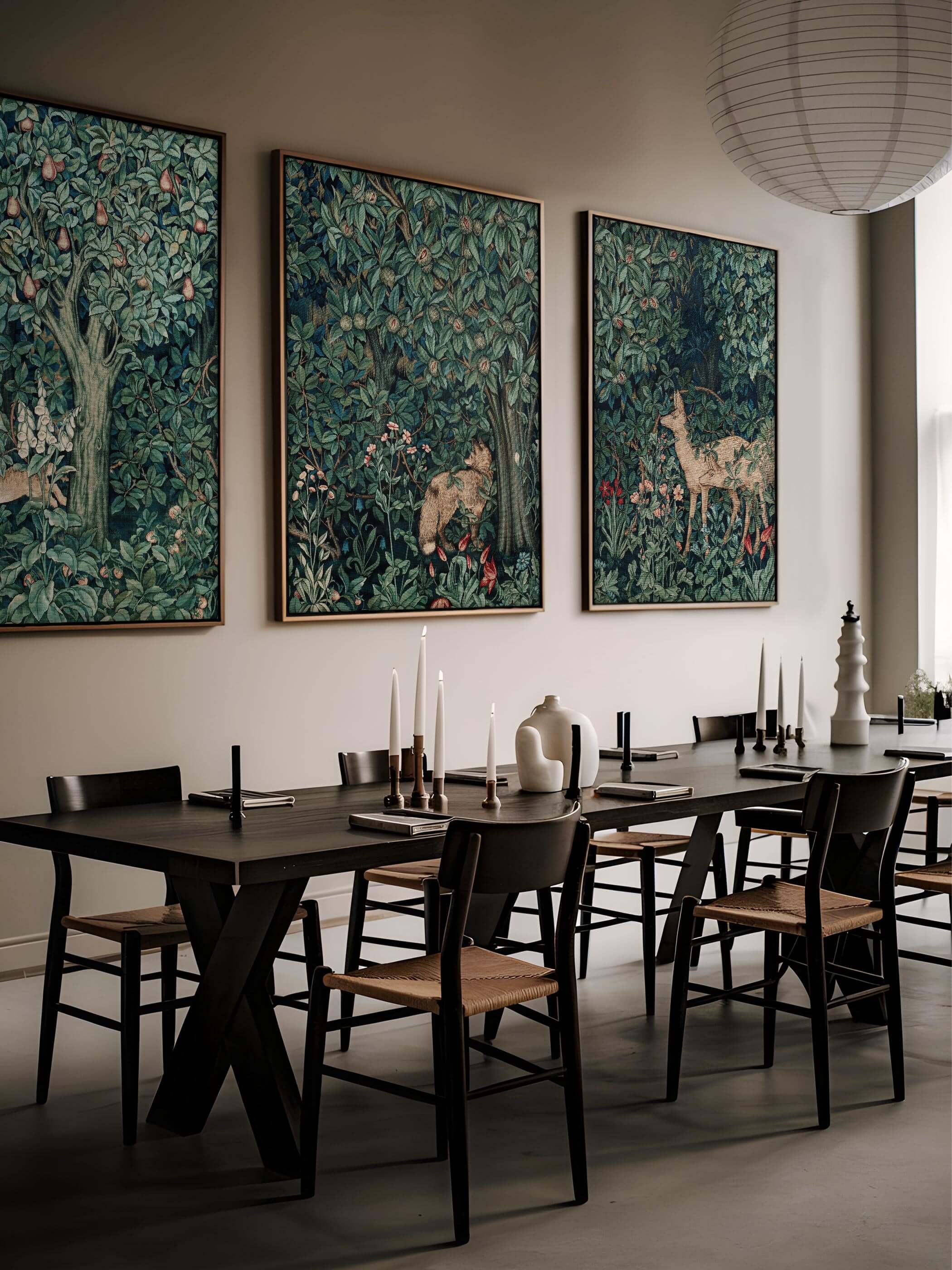 Drei großformatige William Morris Poster im Esszimmer an der Wand hängend. Die detaillierten Waldszenen in tiefen Grüntönen zeigen verschiedene Tiere und Pflanzen. Der minimalistische Esstisch mit schwarzen Stühlen und Kerzenhaltern unterstreicht den eleganten Look des Raumes.