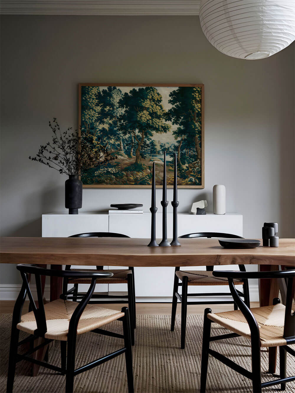 Stilvolles Esszimmer mit einem großen, gerahmten Rokoko-Wandteppich-Druck über einer Kommode und einem minimalistischen Holztisch mit schwarzen Stühlen. Der Druck zeigt einen Wald mit Bäumen und einen See im Hintergrund.