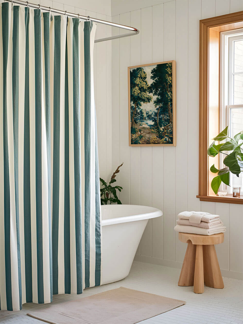 Eine Rokoko-Tapisserie mit malerischer Waldszene in Blau- und Grüntönen, als gerahmtes Poster an der Wand eines hellen Badezimmers mit blau-weißem Duschvorhang und Holzdekor befestigt.