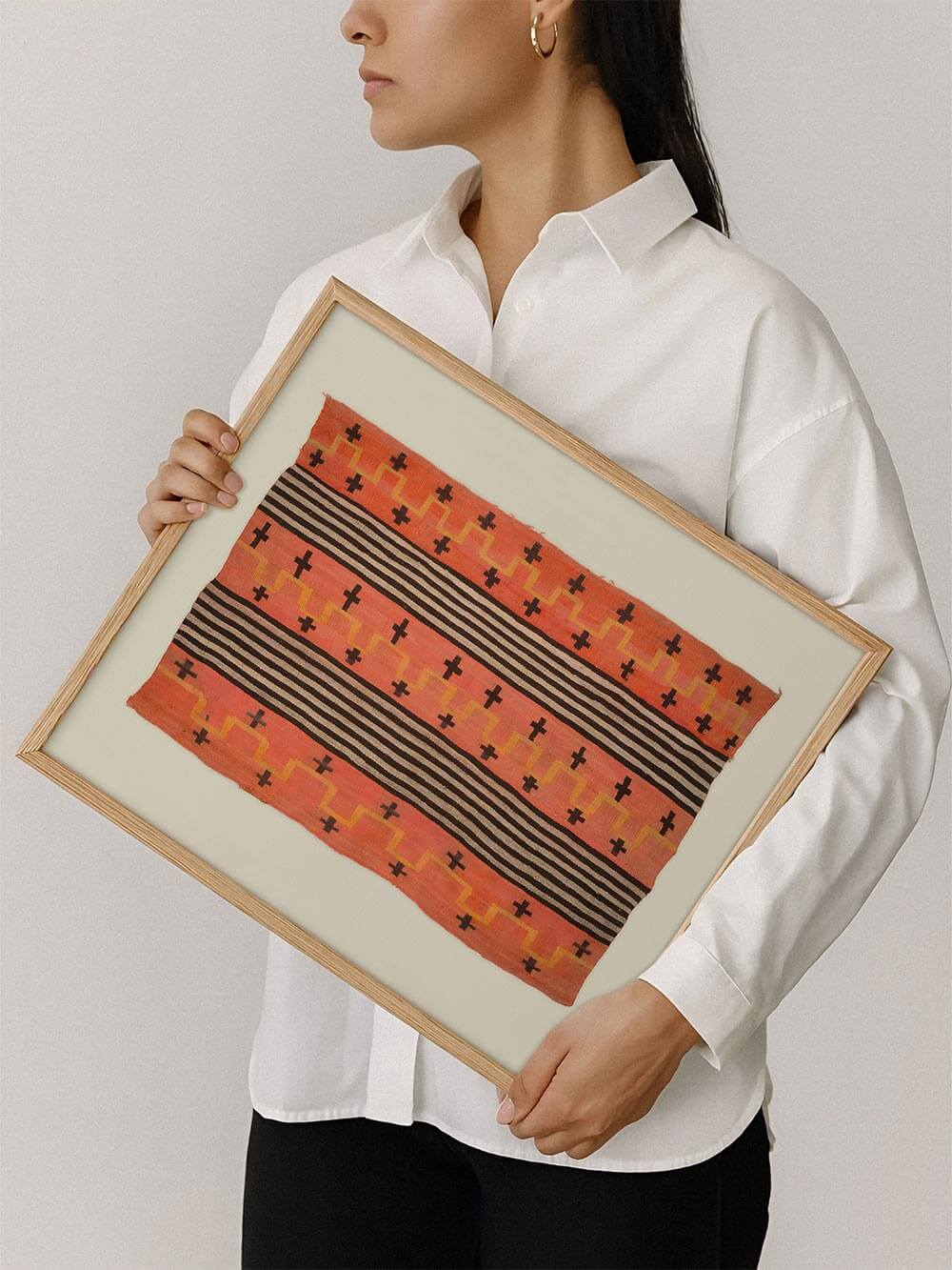 Gerahmter Kunstdruck eines grafischen Textilmotivs in Orange, Schwarz und Beige wird von einer Person gehalten. Die handgewebte Anmutung und klaren Streifen verleihen dem Wandbild eine warme Vintage-Boho-Stimmung und zeigen Größe sowie Wirkung des Posters.