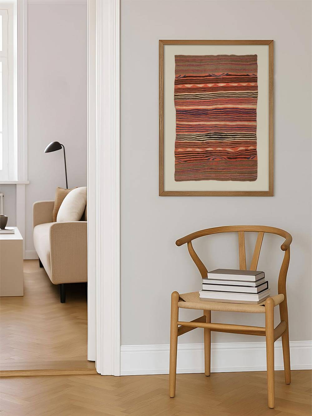 Das textile Wandbild hängt an einer hellen Wand über einem Holzstuhl auf dem gestapelten Büchern liegen. Der Kunstdruck setzt einen warmen, ruhigen Akzent und fügt sich harmonisch in minimalistische Wohnräume oder skandinavisch inspirierte Interieurs ein.