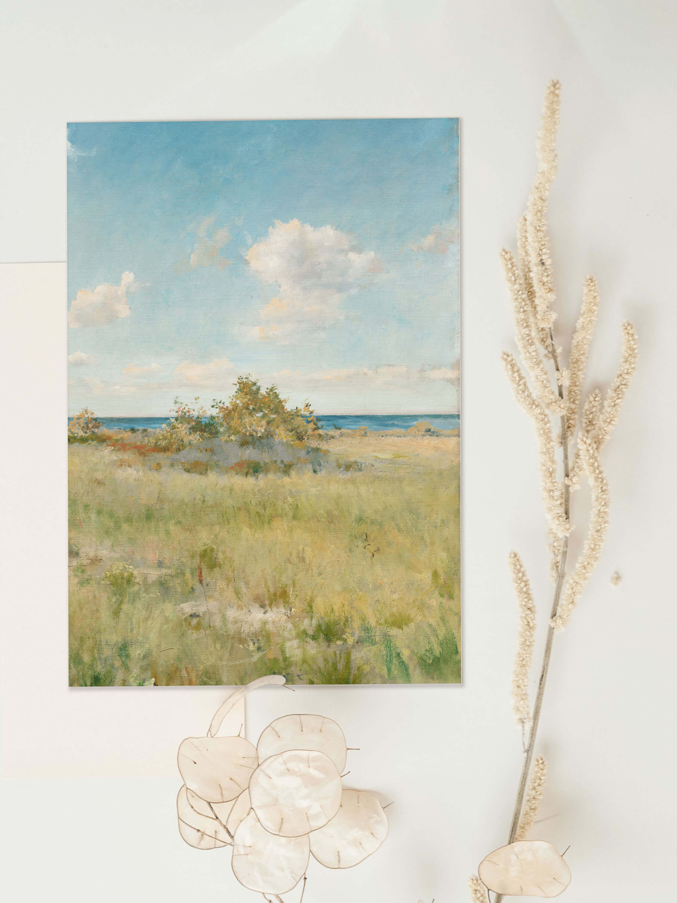 Kunstdruck mit sommerlicher Küstenwiese liegt flach neben getrockneten Gräsern und Silberblatt-Zweigen. Die Nahaufnahme zeigt die Papierqualität und die sanften Pastelltöne des Landschaftsmotivs im Detail.